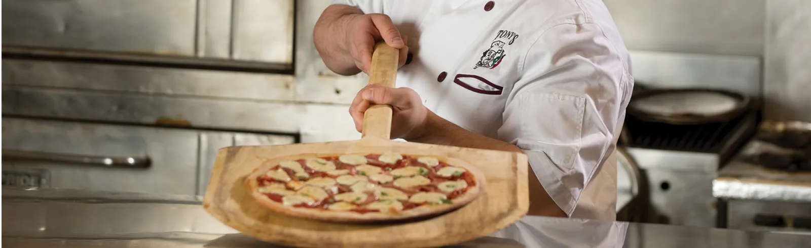 A Tony's pizzaiolo in branded whites sliding a fresh pie from the oven on a wooden peel
